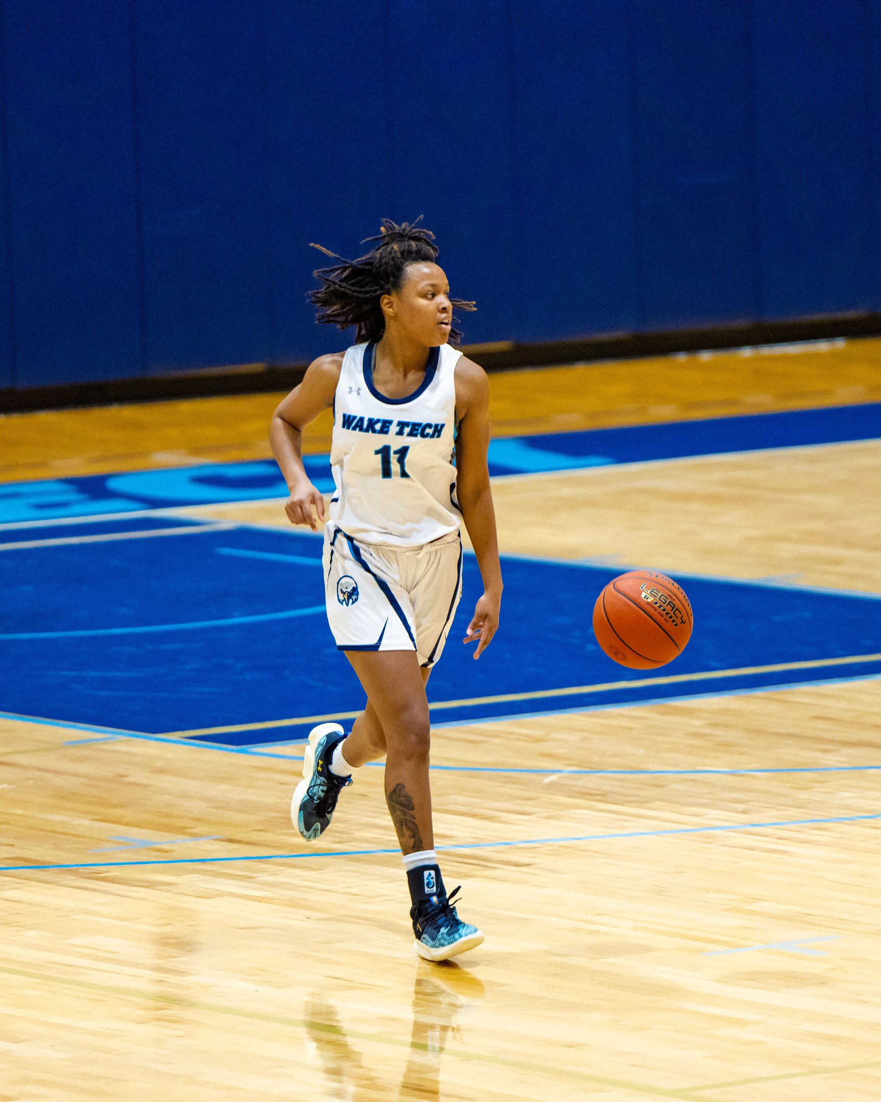 Eine Frau in einer blauen und weißen Basketballuniform, die einen Basketball auf einem Court dribbelt, trägt ein weißes T-Shirt mit der Aufschrift "Wake Tech Women's Basketball" und blaue Schuhe, vor einer blauen Wand.