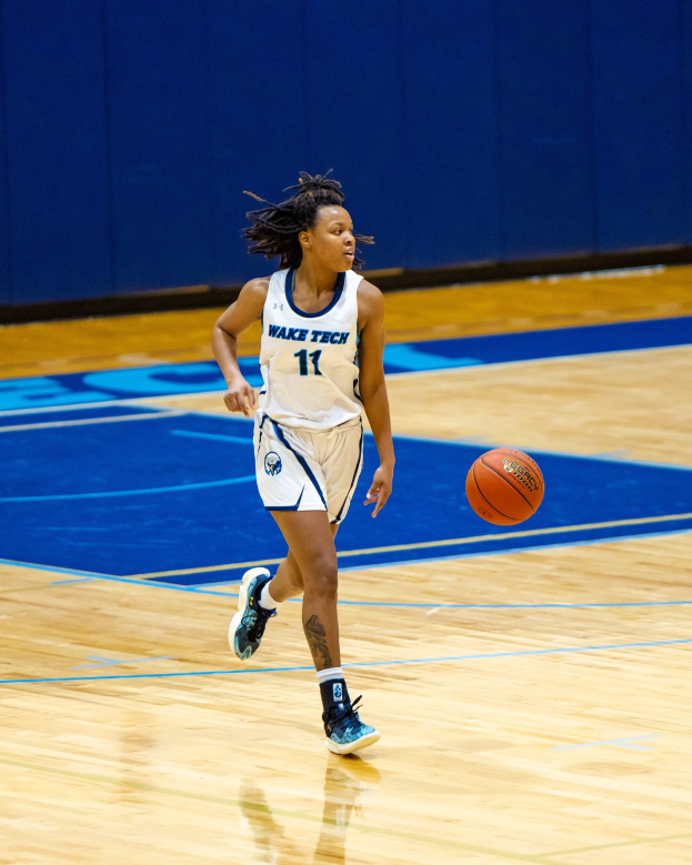 Eine Frau in einer blauen und weißen Basketballuniform, die einen Basketball auf einem Court dribbelt, trägt ein weißes T-Shirt mit der Aufschrift "Wake Tech Women's Basketball" und blaue Schuhe, vor einer blauen Wand.