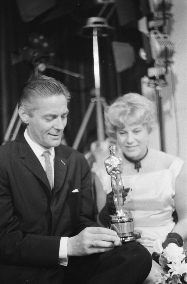 Ein Schwarz-Wei├č-Bild eines formell gekleideten Mannes, der eine Oscar-Statue h├Ąlt und eine Frau, die einen Blumenstra├čus h├Ąlt, nebeneinander sitzend mit Hintergrundbeleuchtung.