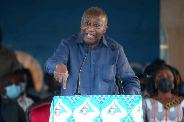 Ein Mann in einem blauen Hemd spricht in zwei Mikrofone an einem Rednerpult, hinter ihm eine Menge und mehrere sitzende Personen, einige tragen Masken.