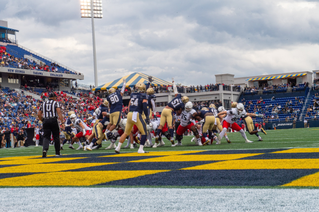 Fuballspiel in einem Stadion mit Spielern in Sportkleidung und Helmen im Vordergrund, Zuschauern im Hintergrund und Text unten.