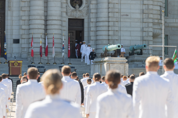 Gruppe von Menschen in weißen Marineuniformen, die auf einer Treppe vor einem Gebäude mit Säulen, Fahnen, einem Podium und Kanonen während einer Abschlussfeier stehen.