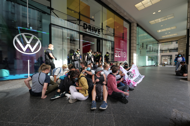 Eine Gruppe von Menschen protestiert vor einer Volkswagen-Niederlassung, hält eine Fahne und setzt sich vor dem gläsernen Gebäude auf den Boden.