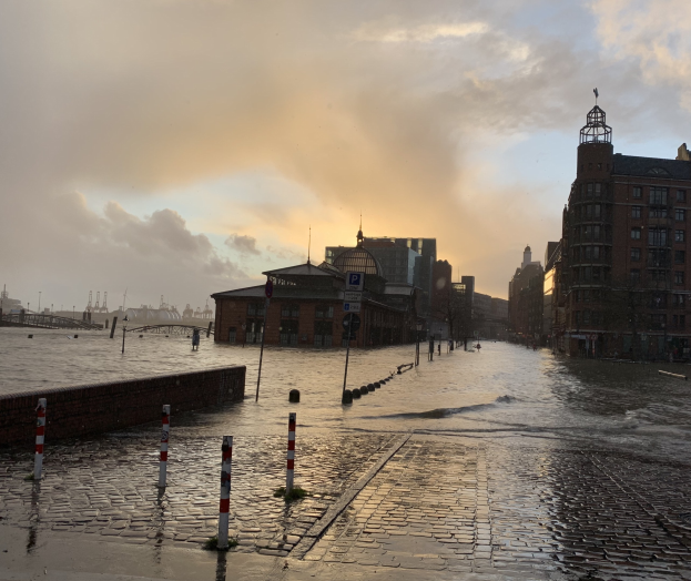 Uberflutete Straße in Hamburg, Deutschland, mit Wasser auf der Straße, Masten, Schildern, Gebäuden mit Fenstern, einer Brücke und einem bewölktem Himmel im Hintergrund.