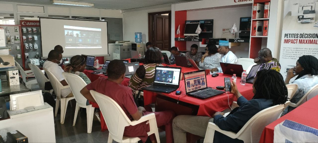 Gruppe von Menschen an einem Tisch mit Laptops, Flaschen und anderen Gegenständen während eines digitalen Marketing-Workshops, mit einem Banner, einem Bildschirm, einer Tür und Deckenlampen im Hintergrund.