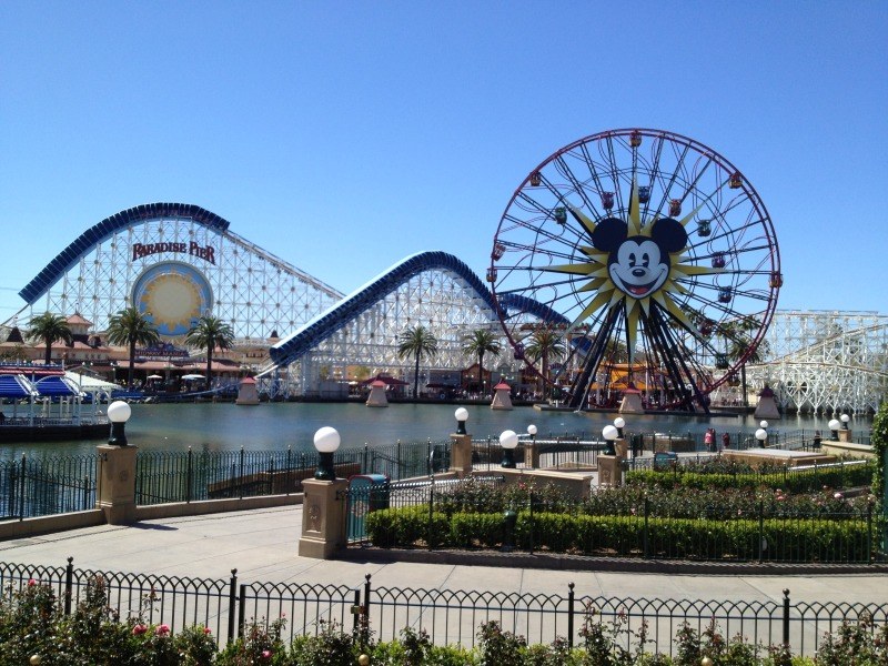Luftaufnahme von Disney California Adventure mit seinem Riesenrad und Achterbahn, umgeben von Grünflächen und Laternenmasten, mit Wasser im Vordergrund unter einem klaren blauen Himmel.