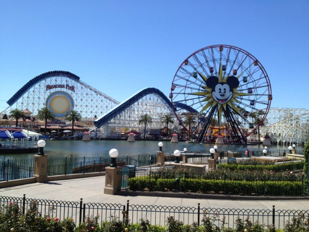 Luftaufnahme von Disney California Adventure mit seinem Riesenrad und Achterbahn, umgeben von Grünflächen und Laternenmasten, mit Wasser im Vordergrund unter einem klaren blauen Himmel.