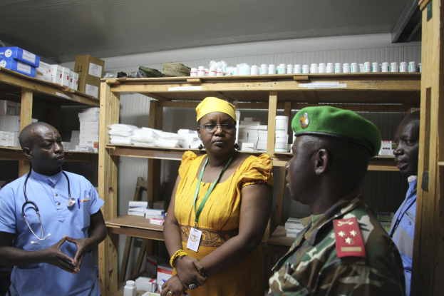 Vier Personen in einer Apotheke, mit einer Frau in einem gelben Kleid und grüner Mütze zwischen zwei Männern in blauen Hemden mit Stethoskopen, umgeben von Medikamentenflaschen und -schachteln.